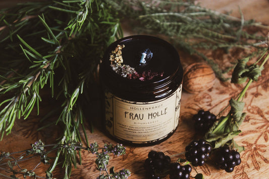 Kräuter-Kerze "Frau Holle" - Kerze - Handgemachte Frau Holle Ritualkerze aus Recyclingwachs in einem dunklen Glasbehälter, umgeben von natürlichen Elementen wie grünen Kräutern, Brombeeren und einer Walnuss auf einer hölzernen Oberfläche. Die Kerze verströmt einen Duft von dichtem Kiefernwald, ergänzt durch Noten von Beifuß, Mohn und Holunder, den Kräutern der Holle. Offene Kerze mit Docht, man sieht ein Topping an Kräutern