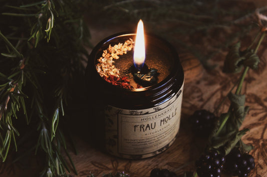 Kräuter-Kerze "Frau Holle" - brennende Kerze - Handgemachte Frau Holle Ritualkerze aus Recyclingwachs in einem dunklen Glasbehälter, umgeben von natürlichen Elementen wie grünen Kräutern, Brombeeren und einer Walnuss auf einer hölzernen Oberfläche. Die Kerze verströmt einen Duft von dichtem Kiefernwald, ergänzt durch Noten von Beifuß, Mohn und Holunder, den Kräutern der Holle.