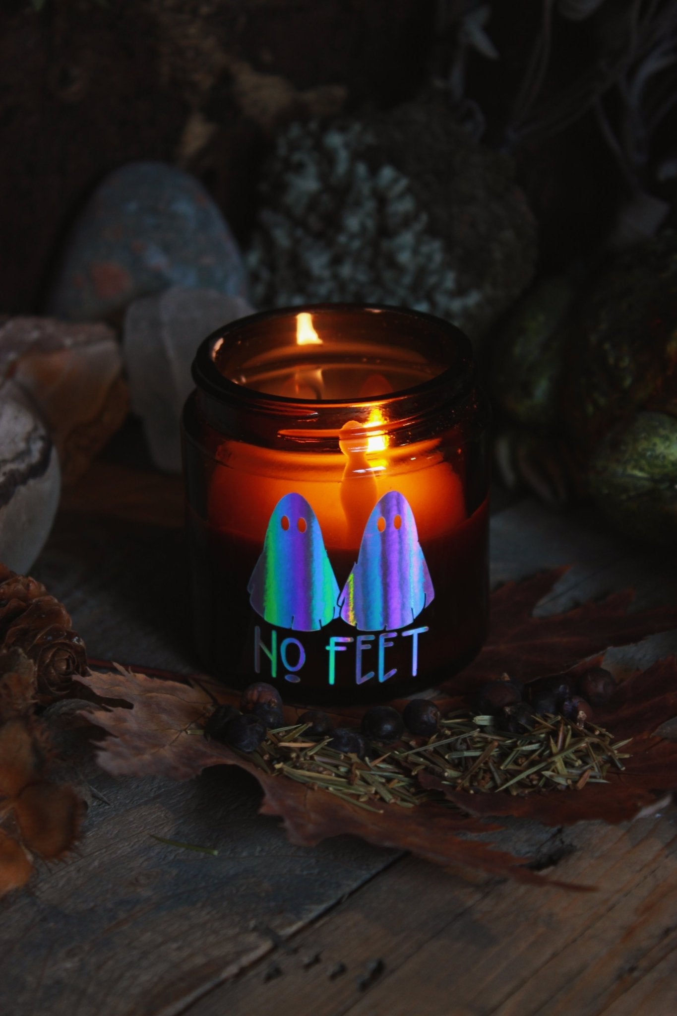 Kräuter-Kerze "No Feet" - Das Bild zeigt ein atmosphärisches Bild eines brennenden Kerzenglases mit dem Aufdruck “NO FEET” und zwei bunten Geisterbildern, umgeben von herbstlichen Elementen wie trockenen Blättern und Tannenzapfen.
