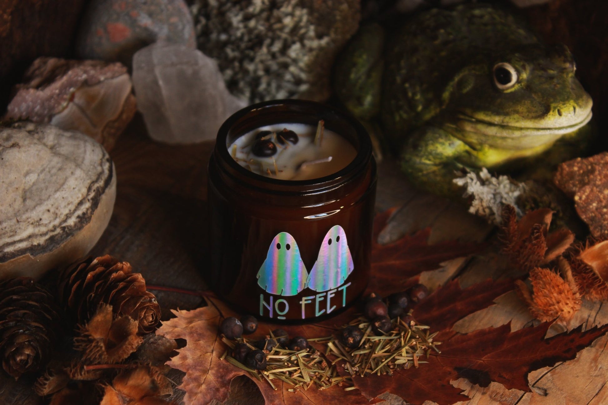 Kräuter-Kerze "No Feet" - Das Bild zeigt ein atmosphärisches Bild eines Kerzenglases mit dem Aufdruck “NO FEET” und zwei bunten Geisterbildern, umgeben von herbstlichen Elementen wie trockenen Blättern und Tannenzapfen.