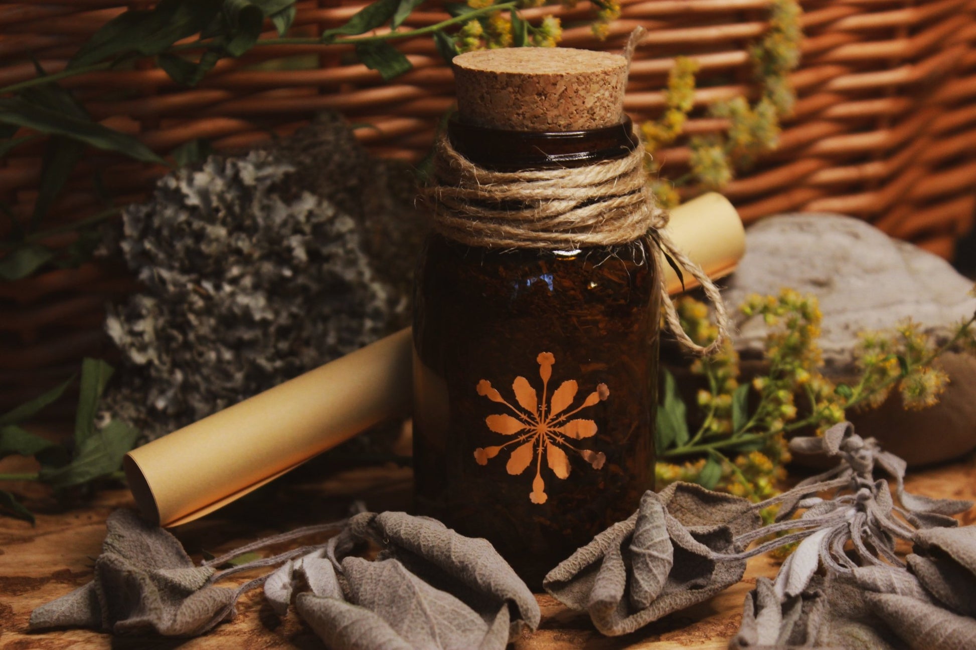 Räucherwerk Trotula -Kleines braunes Glasfläschchen mit Korken und einem floralen Logo, umwickelt mit einer Schnur und dekoriert mit getrockneten Blättern, präsentiert vor einem geflochtenen Korb, umgeben von gelben Blüten und rustikalen Elementen. 