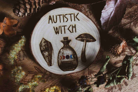 Nahaufnahme Wandbild aus Holz "Autistic Witch" - Handgefertigte Holzgravur mit dem Text ‘AUTISTIC WITCH’, umgeben von detaillierten Zeichnungen eines Blattes, einer Flasche mit farbiger Flüssigkeit und eines Pilzes. alles ist umgeben von Kräutern und Pflanzen