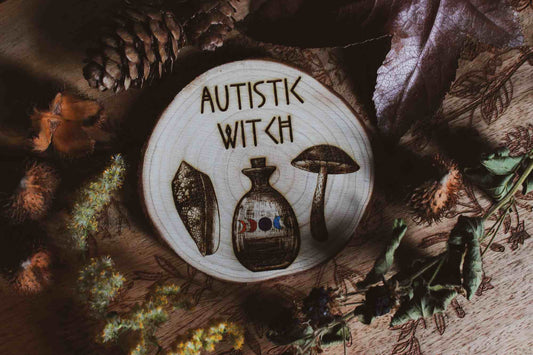 Wandbild aus Holz "Autistic Witch" - Handgefertigte Holzgravur mit dem Text ‘AUTISTIC WITCH’, umgeben von detaillierten Zeichnungen eines Blattes, einer Flasche mit farbiger Flüssigkeit und eines Pilzes. alles ist umgeben von Kräutern und Pflanzen