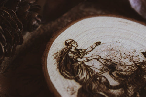 Nahaufnahme Wandbild aus Holz "Freyja im Katzenwagen" - Wandbild aus Holz "Freyja im Katzenwagen" - “Handgefertigtes Kunstwerk auf einem kreisförmigen Holzstück, das eine Freya auf einem Zugwagen darstellt, Katzen ziehen ihn Das Kunstwerk ist umgeben von Naturmaterialien wie Tannenzapfen und Blättern. 