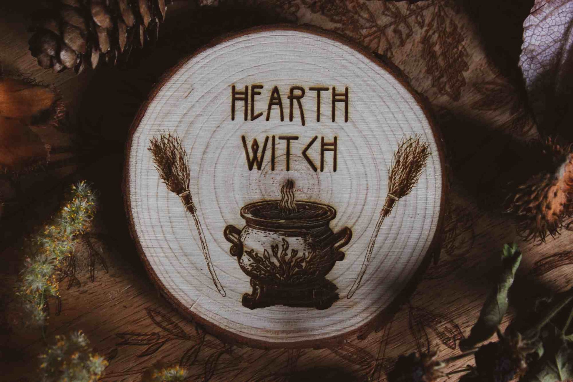 Wandbild aus Holz "Hearth Witch" -Bild eines hölzernen Gegenstands mit der Inschrift ‘Hearth Witch’, auf der Scheibe sieht man 2 Besen die einen Hexenkessel umrahmen umgeben von natürlichen Elementen wie Tannenzapfen und Blumen, 