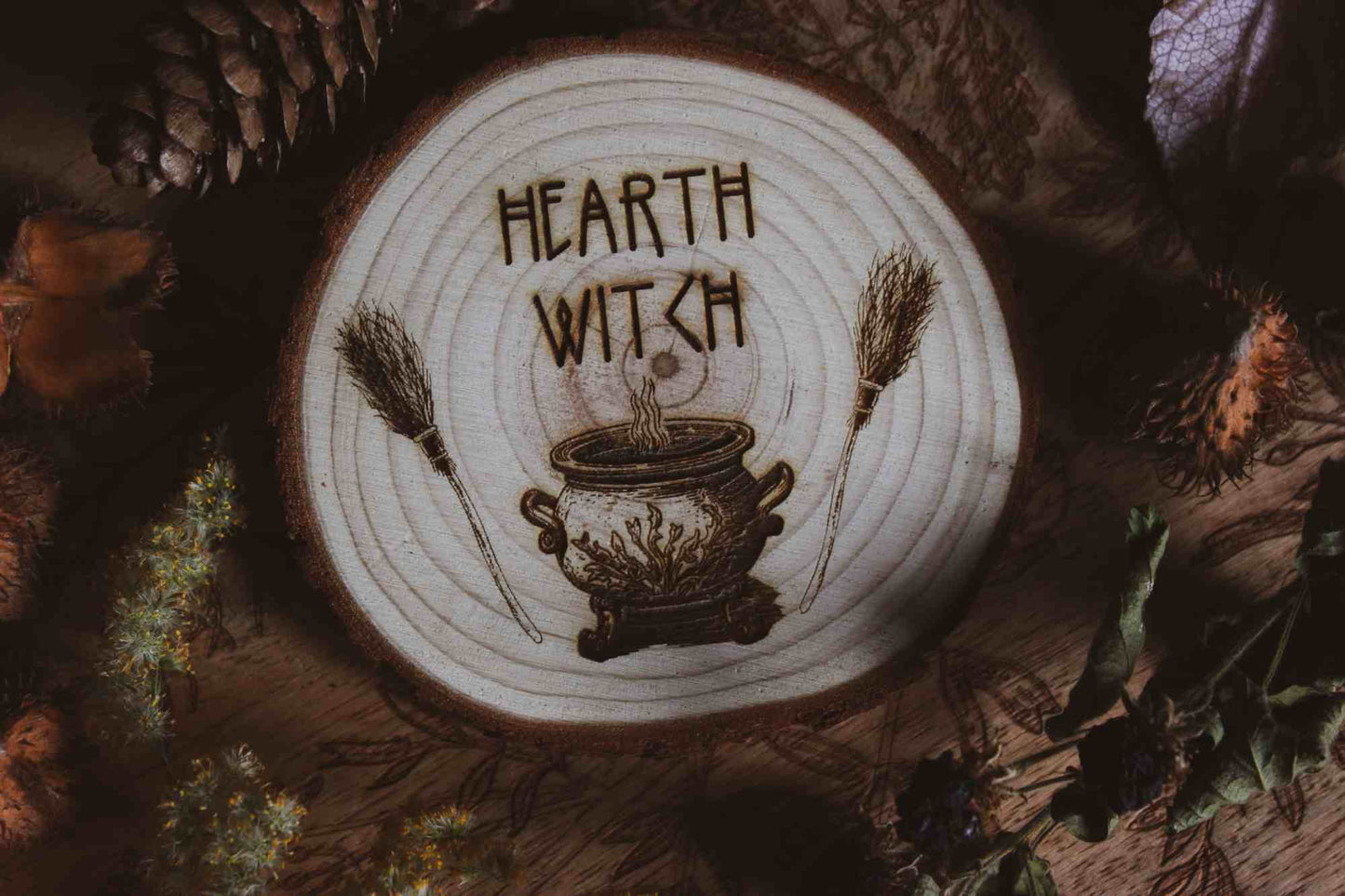 Wandbild aus Holz "Hearth Witch" -Bild eines hölzernen Gegenstands mit der Inschrift ‘Hearth Witch’, auf der Scheibe sieht man 2 Besen die einen Hexenkessel umrahmen umgeben von natürlichen Elementen wie Tannenzapfen und Blumen, 