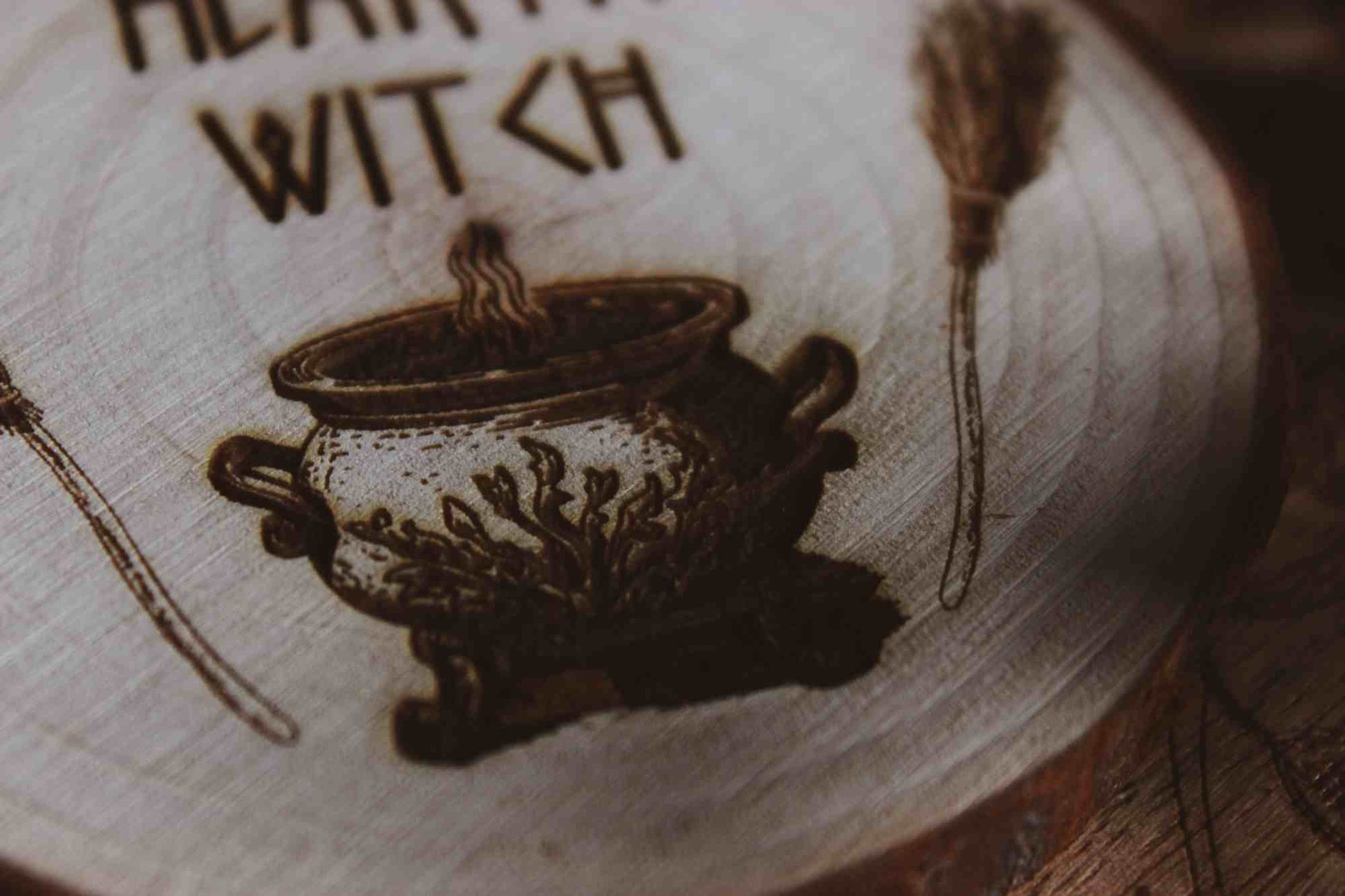 Nahaufnahme Wandbild aus Holz "Hearth Witch" - Wandbild aus Holz "Hearth Witch" -Bild eines hölzernen Gegenstands mit der Inschrift ‘Hearth Witch’, auf der Scheibe sieht man 2 Besen die einen Hexenkessel umrahmen u