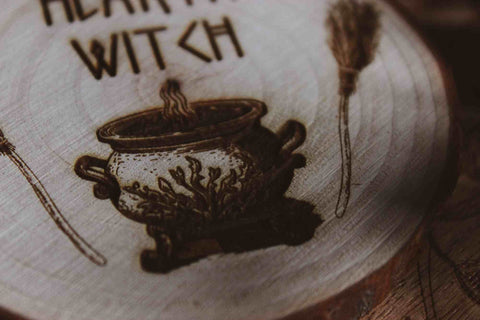 Nahaufnahme Wandbild aus Holz "Hearth Witch" - Wandbild aus Holz "Hearth Witch" -Bild eines hölzernen Gegenstands mit der Inschrift ‘Hearth Witch’, auf der Scheibe sieht man 2 Besen die einen Hexenkessel umrahmen u