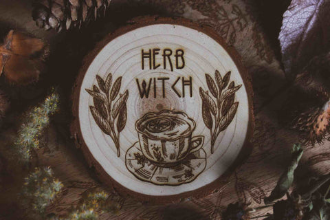 Wandbild aus Holz "Herb Witch" - Dieses Bild zeigt eine kunstvoll gravierte Holzscheibe mit der Inschrift “HERB WITCH”, umgeben von einer natürlichen und mystischen Atmosphäre. Die Gravur stellt eine Tasse Tee dar, die von zwei Zweigen mit Blättern flankiert wird, 