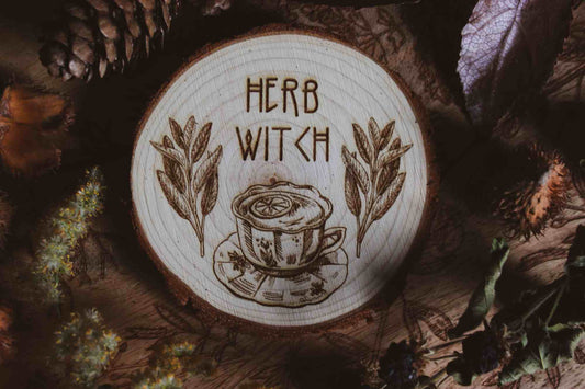 Wandbild aus Holz "Herb Witch" - Wandbild aus Holz "Herb Witch" - Dieses Bild zeigt eine kunstvoll gravierte Holzscheibe mit der Inschrift “HERB WITCH”, umgeben von einer natürlichen und mystischen Atmosphäre. Die Gravur stellt eine Tasse Tee dar, die von zwei Zweigen mit Blättern flankiert wird, 