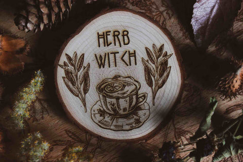 Wandbild aus Holz "Herb Witch" - Dieses Bild zeigt eine kunstvoll gravierte Holzscheibe mit der Inschrift “HERB WITCH”, umgeben von einer natürlichen und mystischen Atmosphäre. Die Gravur stellt eine Tasse Tee dar, die von zwei Zweigen mit Blättern flankiert wird, 