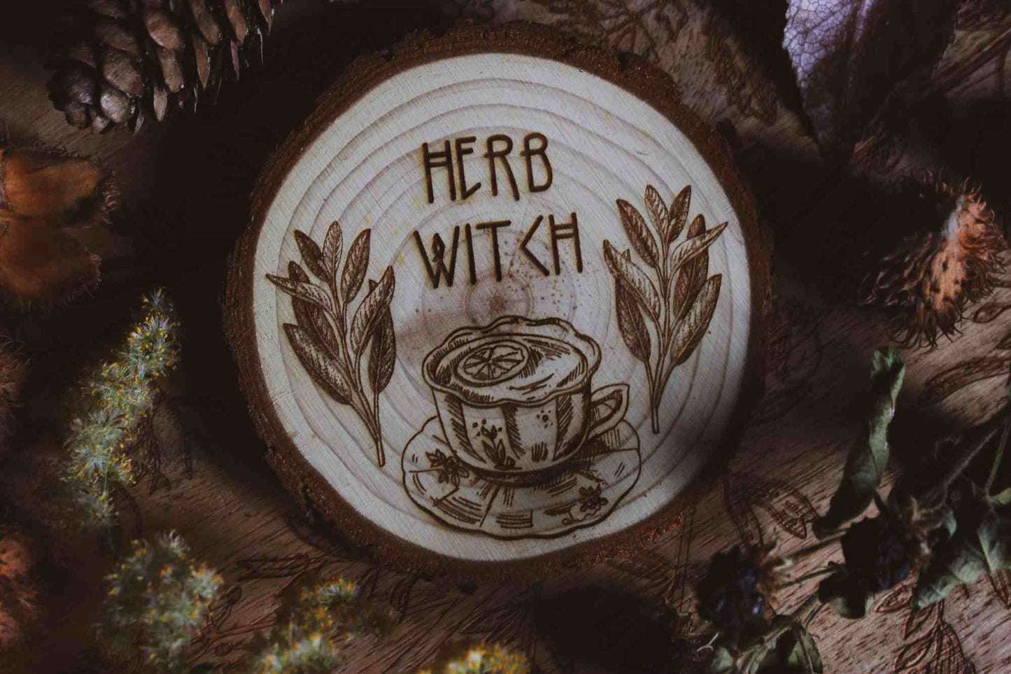 Wandbild aus Holz "Herb Witch" - Dieses Bild zeigt eine kunstvoll gravierte Holzscheibe mit der Inschrift “HERB WITCH”, umgeben von einer natürlichen und mystischen Atmosphäre. Die Gravur stellt eine Tasse Tee dar, die von zwei Zweigen mit Blättern flankiert wird, 