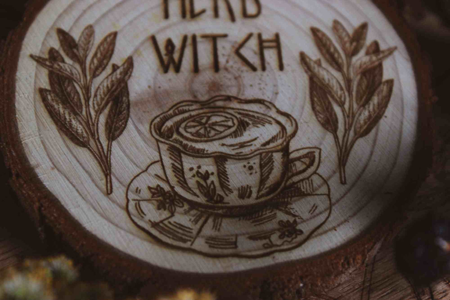 Nahaufnahme Wandbild aus Holz "Herb Witch" - Wandbild aus Holz "Herb Witch" - Dieses Bild zeigt eine kunstvoll gravierte Holzscheibe mit der Inschrift “HERB WITCH”, umgeben von einer natürlichen und mystischen Atmosphäre. Die Gravur stellt eine Tasse Tee dar, die von zwei Zweigen mit Blättern flankiert wird, 