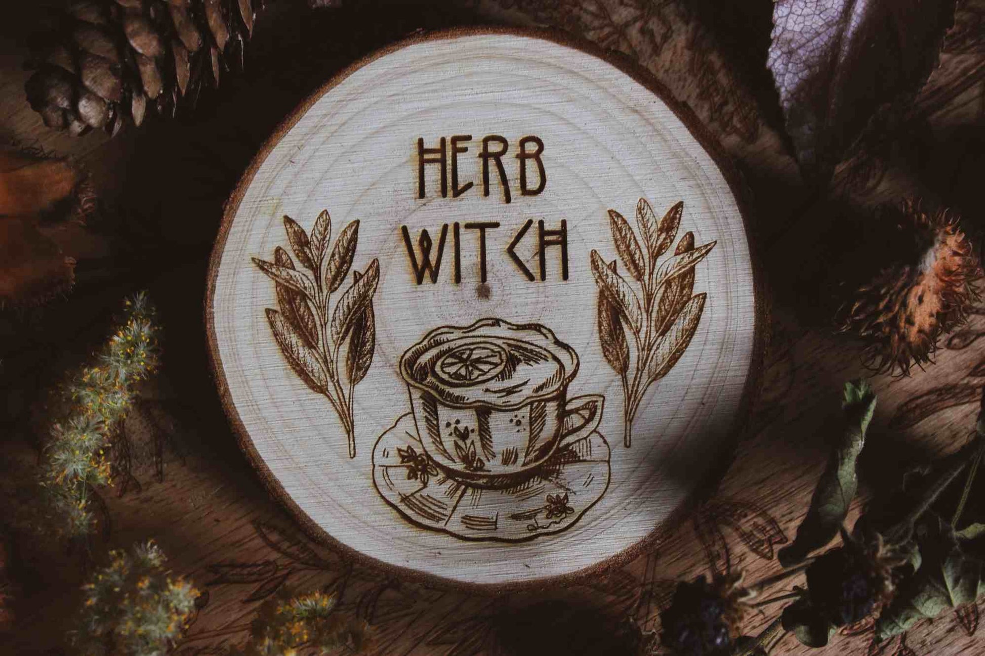 Wandbild aus Holz "Herb Witch" - Dieses Bild zeigt eine kunstvoll gravierte Holzscheibe mit der Inschrift “HERB WITCH”, umgeben von einer natürlichen und mystischen Atmosphäre. Die Gravur stellt eine Tasse Tee dar, die von zwei Zweigen mit Blättern flankiert wird, 