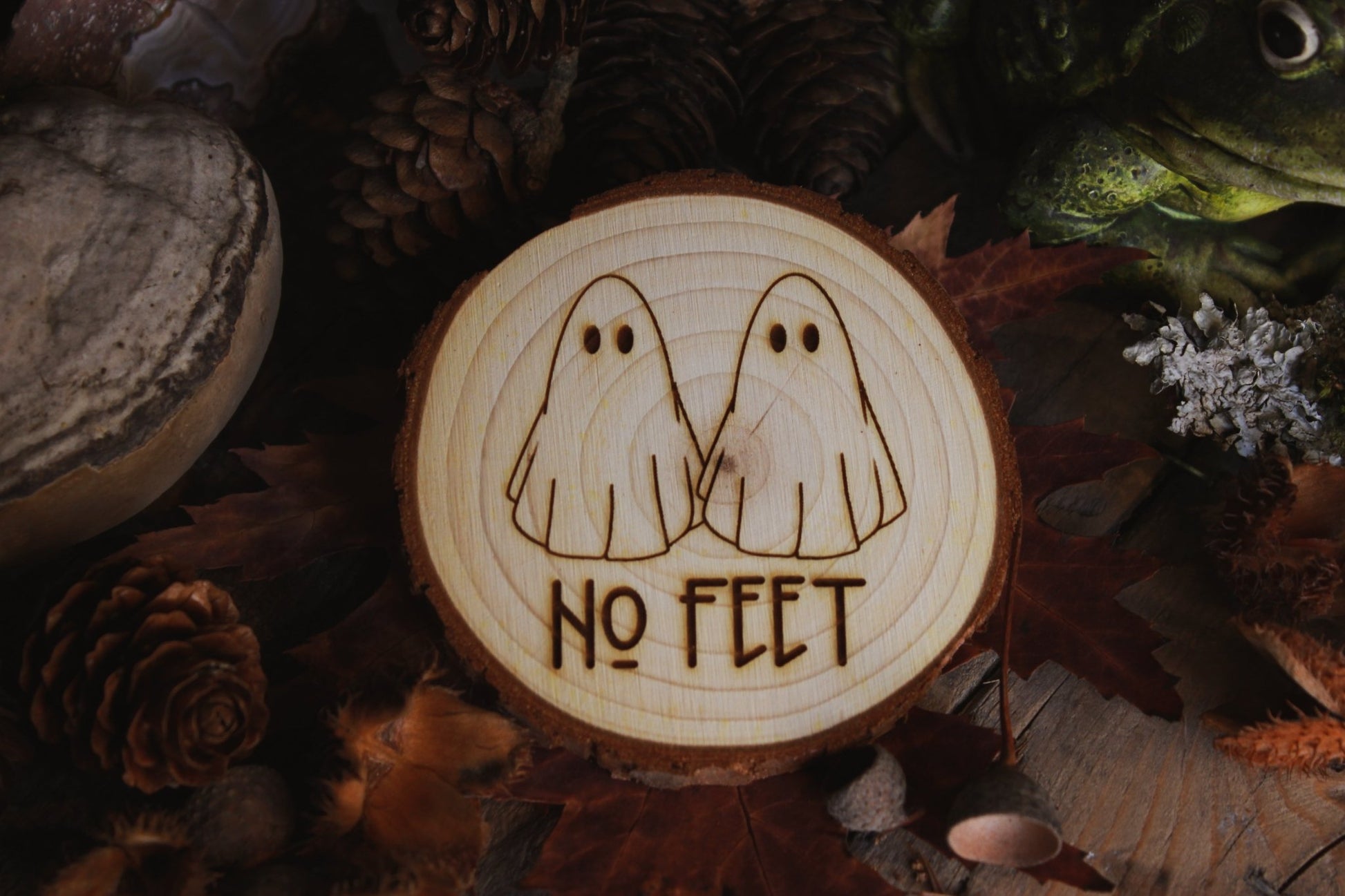  Wandbild aus Holz "No Feet" - Handgefertigtes Holzstück mit einer eingravierten Illustration von zwei Geistern und dem Text ‘NO FEET’, umgeben von natürlichen Elementen wie Tannenzapfen, Steinen und einer Froschstatue.