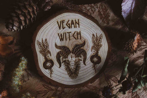 Wandbild aus Holz "Vegan Witch" - Das Bild zeigt ein handgefertigtes Kunstwerk mit der Inschrift “VEGAN WITCH”, umgeben von einer dunklen, mystischen Atmosphäre und natürlichen Elementen wie Tannenzapfen und Blättern.