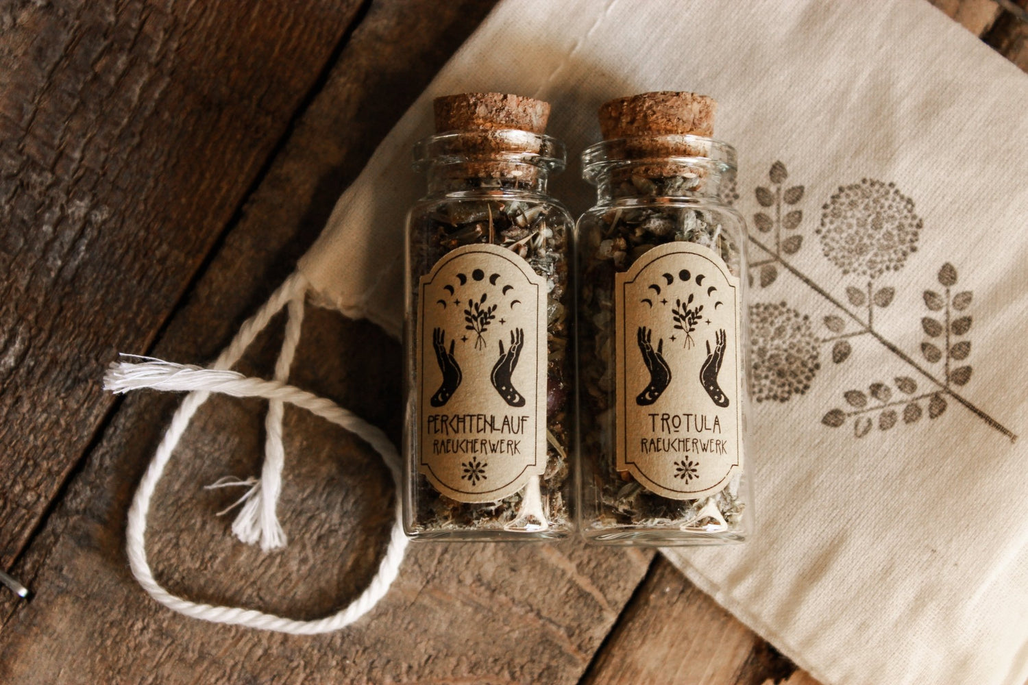 Zwei Glasflaschen ‘Perchtenlauf’ und ‘Trotula’, gefüllt mit getrockneten Kräutern, liegen auf einem rustikalen Holztisch neben einem Stück Stoff mit Blumenmuster.